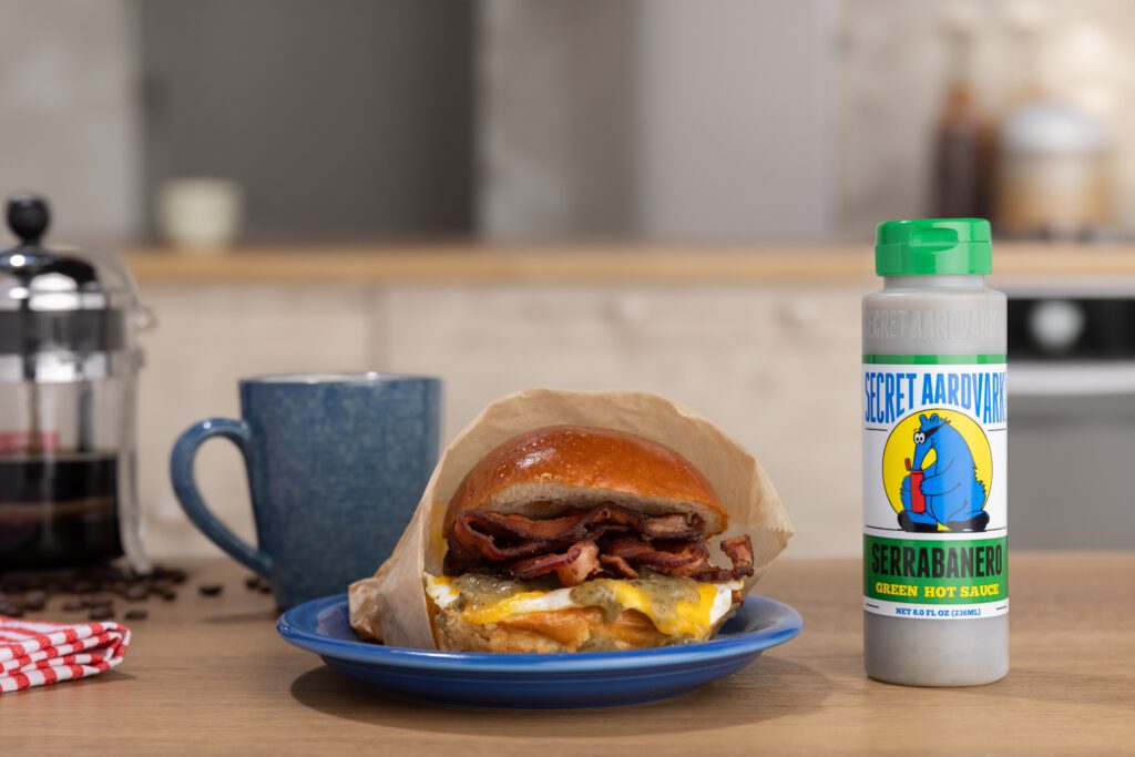 Kitchen counter with french press, coffee mug, a plate with an egg sandwich and bottle of Serrabaner Green Hot Sauce.