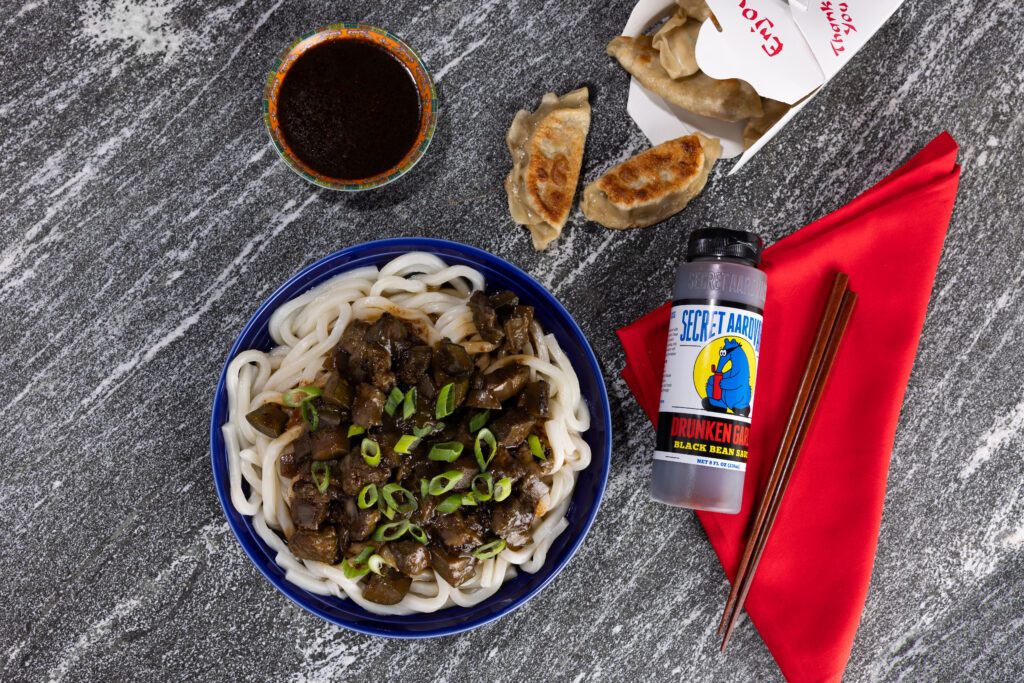 A bowl of noodles with dark protein and green onions on top. Pot stickers coming out of a take out box next to chop sticks and bottle of Drunken Garlic Black Bean Sauce on a red napkin.