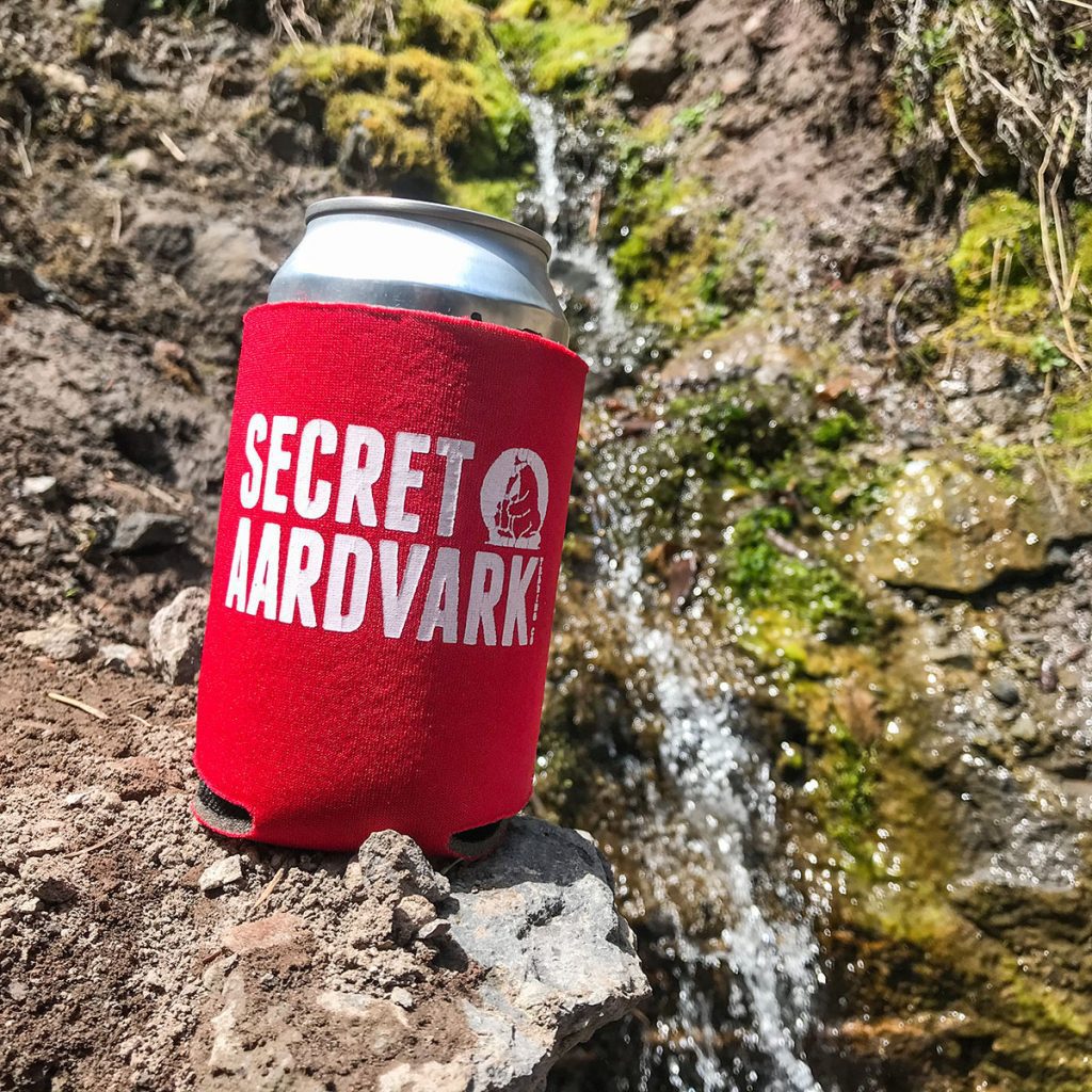 Can of drink in a red secret aardvark koozie sitting on a rock near a stream