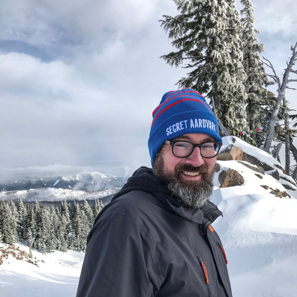 Man smiling and standing on a snowy mountain while wearing a Secret Aardvark beanie