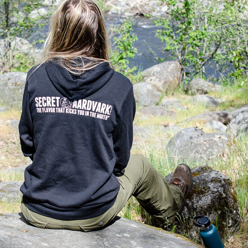 Woman outdoors sitting on a rock near a river wearing a black secret aardvark hoodie
