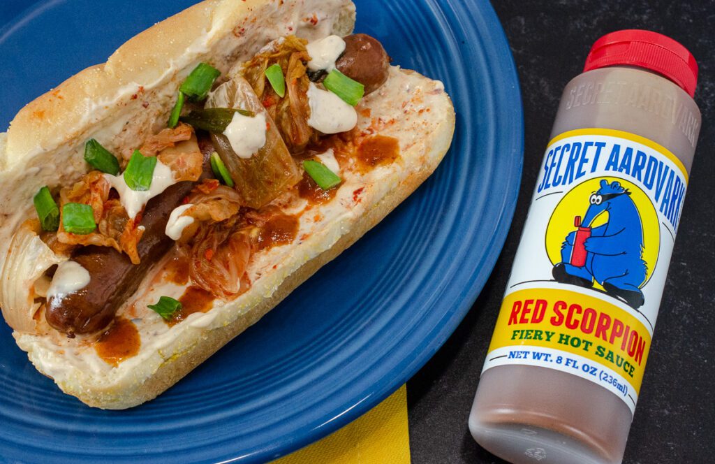 Kimchi hotdog on a plate next to a bottle of Red Scorpion Fiery Hot Sauce