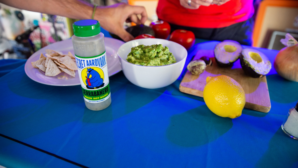 Blue table with avocados, lemon, bottle of Serrbanero and person making guacamole