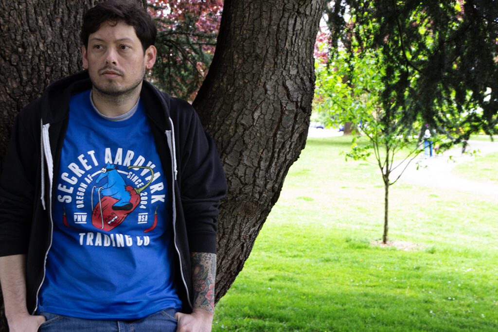 Man leaning against tree with blue Secret Aardvark t-shirt