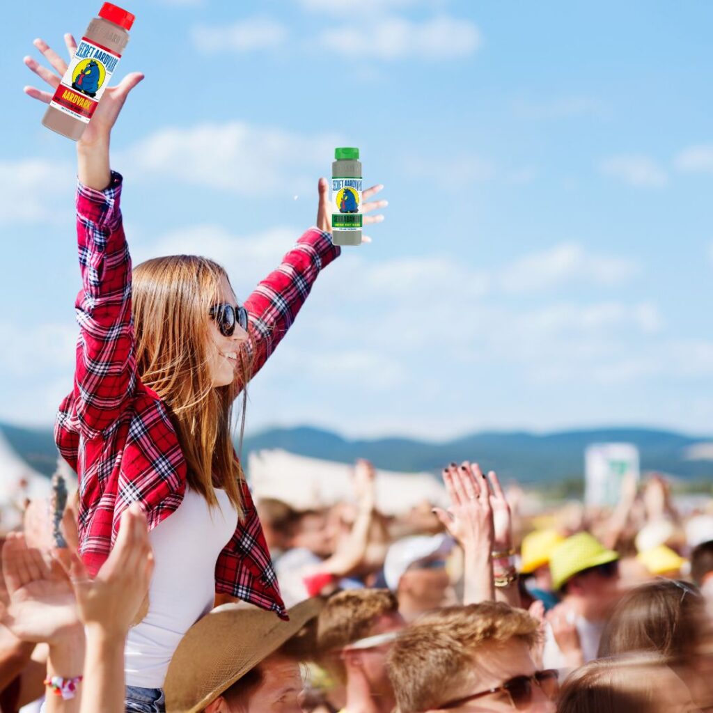 A young female sitting on top of someone's shoulders in a festival crowd holding up bottles of Secret Aardvark hot sauce. 