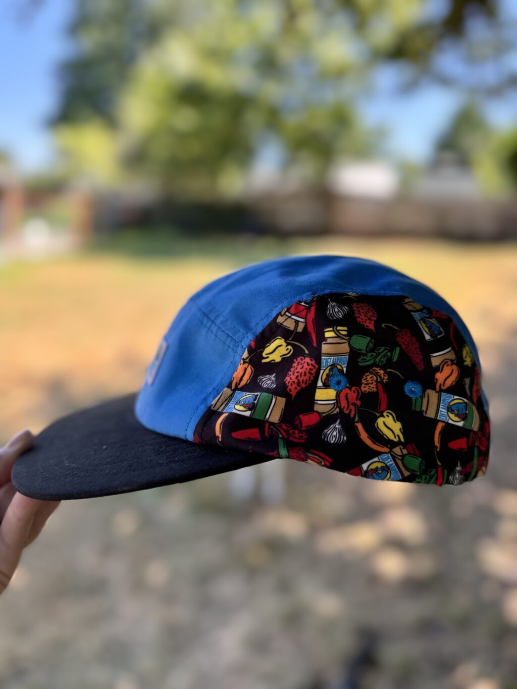 side profile of new side panel camper hat. Hat is blue in the middle, black bill and side panel fabric is bottles of secret aardvark artwork with peppers
