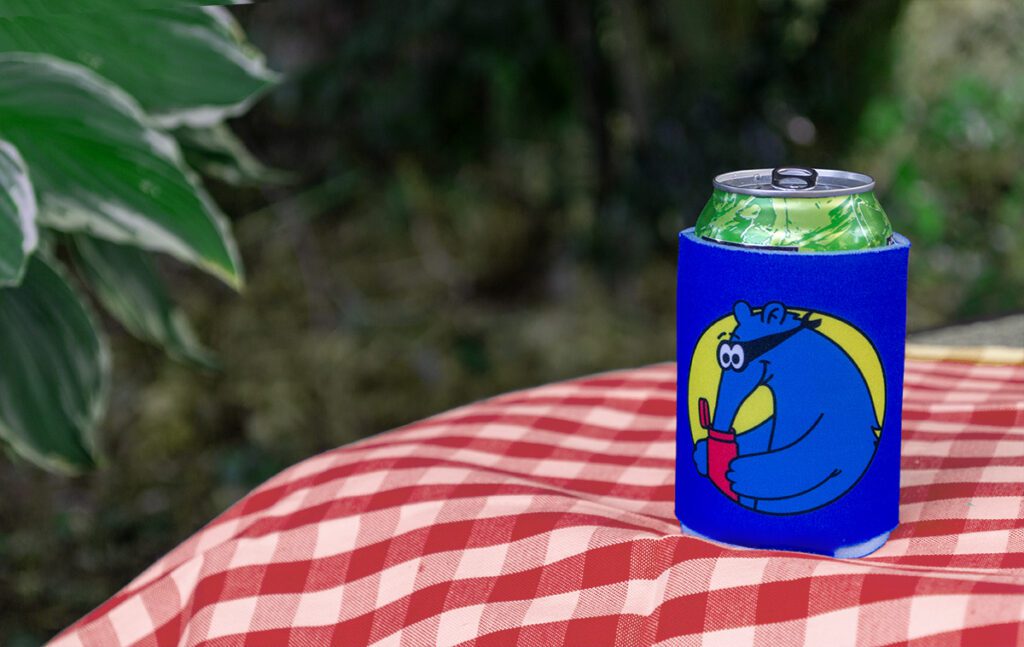 Blue Aardvark Koozie on red picnic table with greenery in the background.