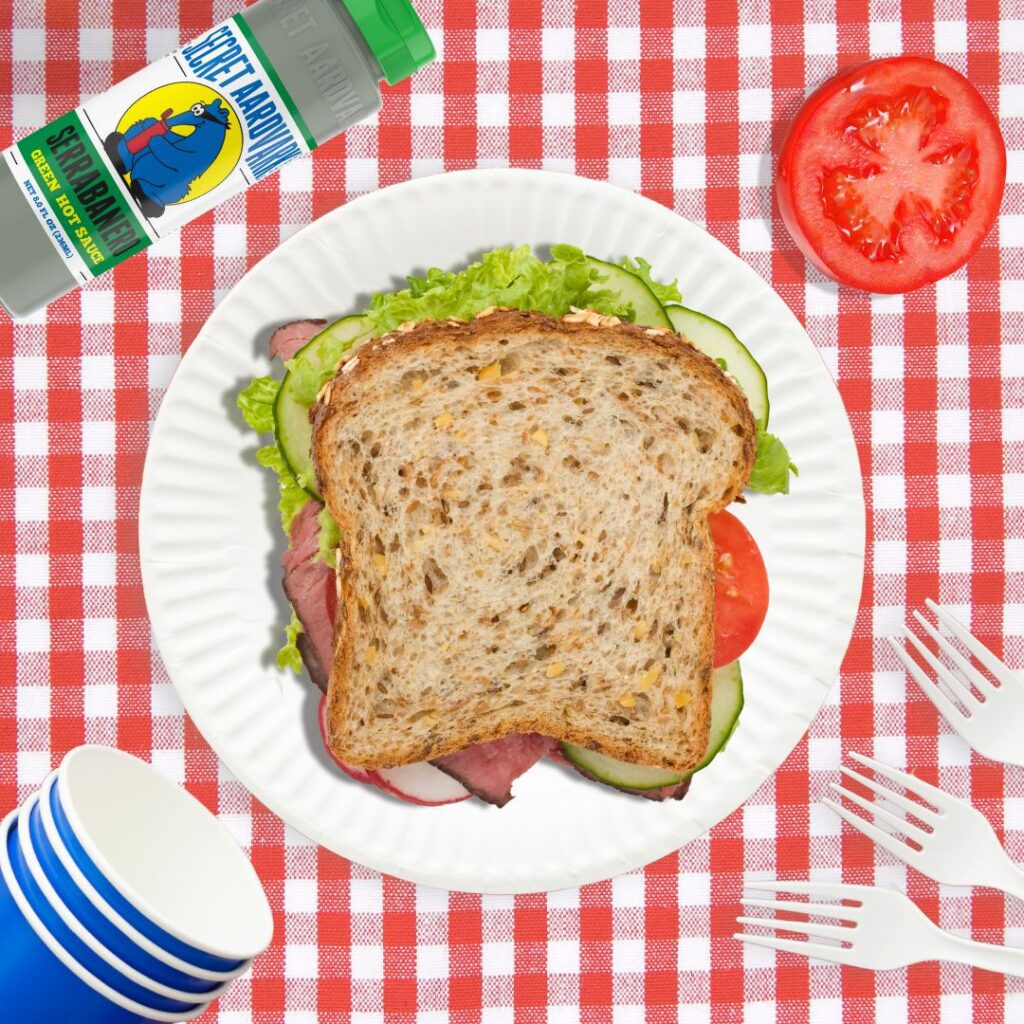 A sandwich on a paper plate on top of a red and white checkered tablecloth.