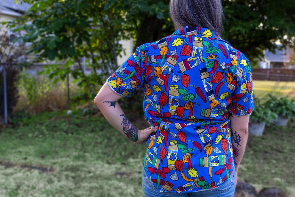 Women standing backwards wearing the new Secret Aardvark button up shirt. The shirt is ble with bright colored peppers and bottles of secret aardvark.