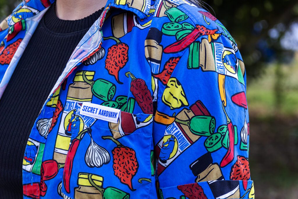 Close up of the new Secret Aardvark button up shirt. The shirt is ble with bright colored peppers and bottles of secret aardvark.