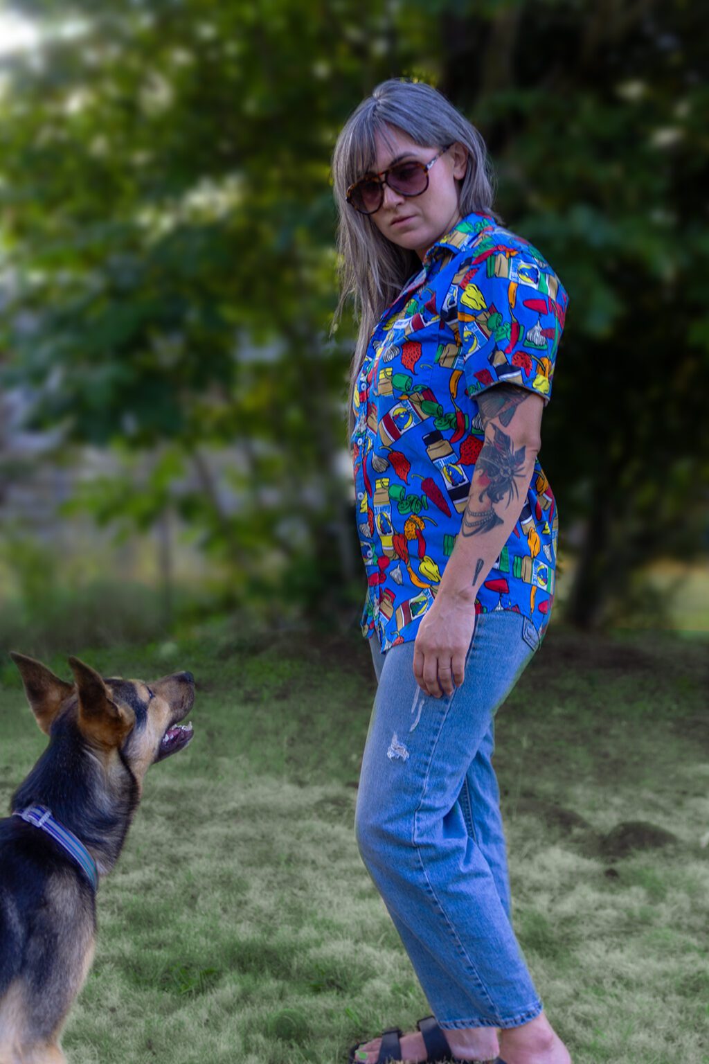 woman standing next to dog, wearing the new Secret Aardvark button up shirt. The shirt is ble with bright colored peppers and bottles of secret aardvark.