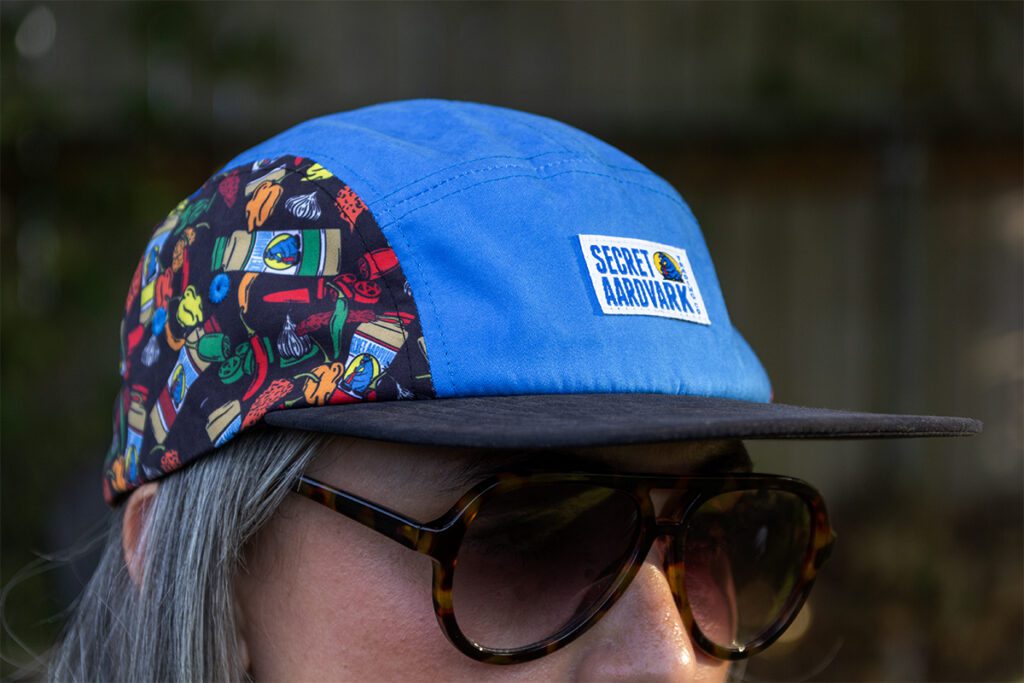 Blue Aardvark camper hat on female wearing sunglasses. Hat is blue in center, with logo patch and side panels of bottles of aardvark and peppers.