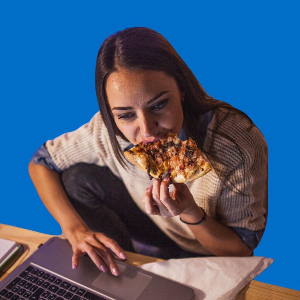 Woman at a desk doing homework while chowing down a slice of pizza.