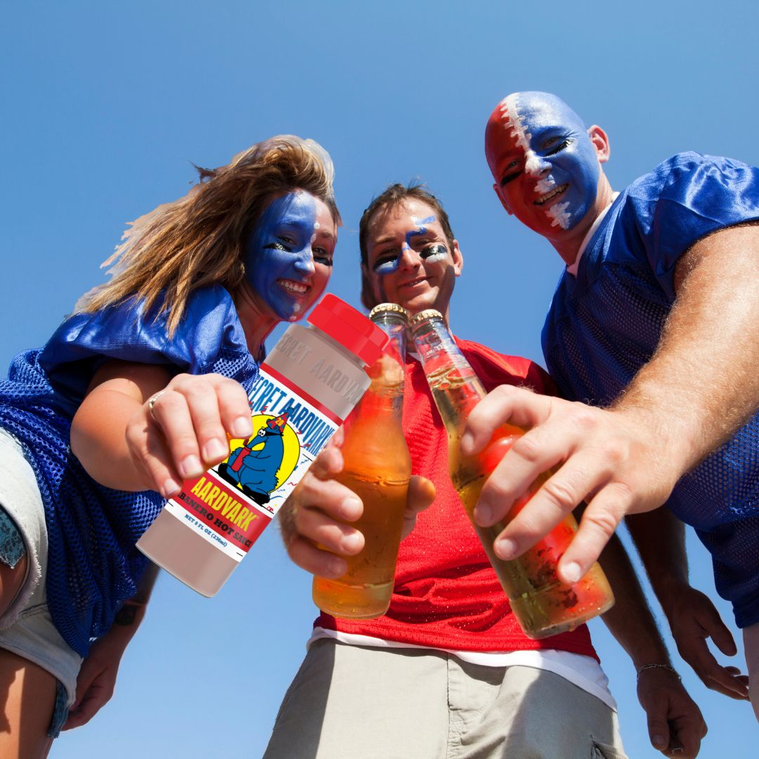 Three people with their faces painted hold beers and hot sauce while the tailgate before a football game.