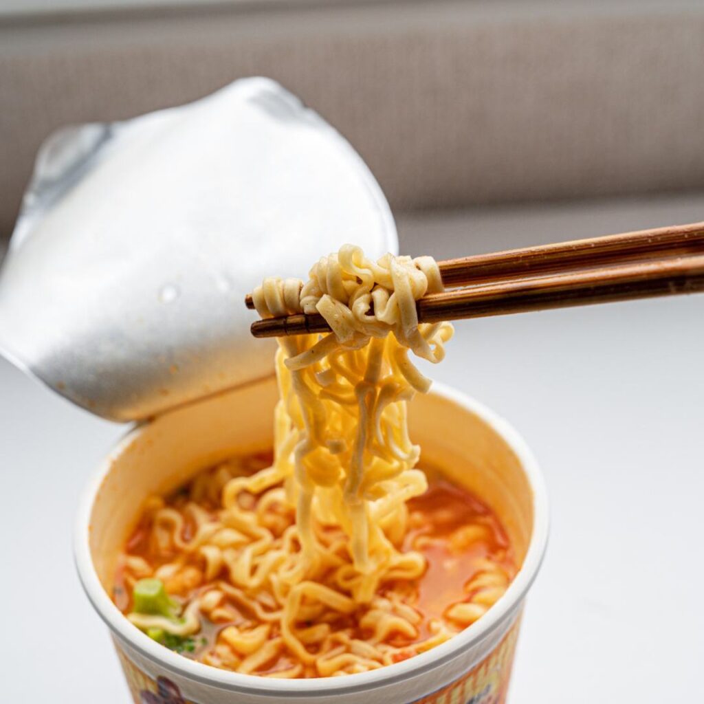 A cup of supermarket ramen noodles. The paper lid is rolled back, noodles are good and hot, and twirled around a pair of chopsticks, ready for that first bite.