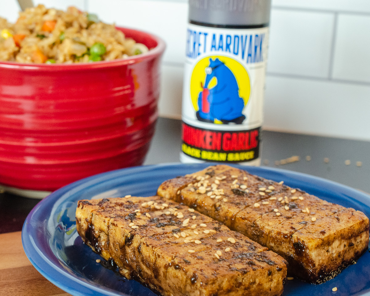 Delicious marinated and cooked tofu slabs cut into an accordion shape, with a red bowl full of fried rice and a bottle of Secret Aardvark Drunken Garlic Black Bean Marinade.