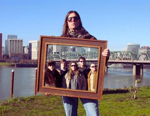 Scott Moritz stands on the Portland, Oregon waterfront holding up a framed portrait of his band the Slow Drags.